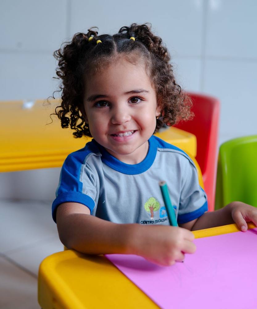 Aluna do CES, de uniforme, sorrindo enquanto desenha na sala de aula